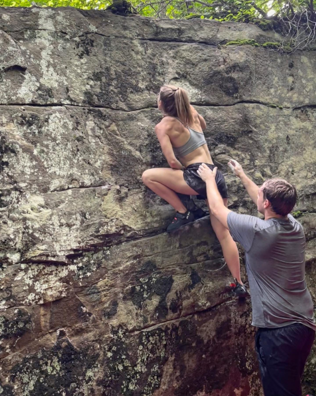 Woman rock climbing outside with person spotting her from below.