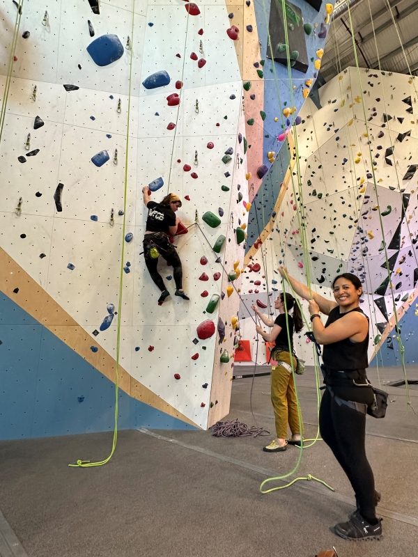 Woman smiling at camera while belaying climbing partner at indoor rock gym.