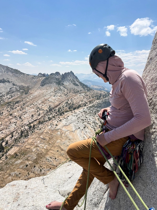 Rock climber belaying on cliff edge with mountain scape in the background.