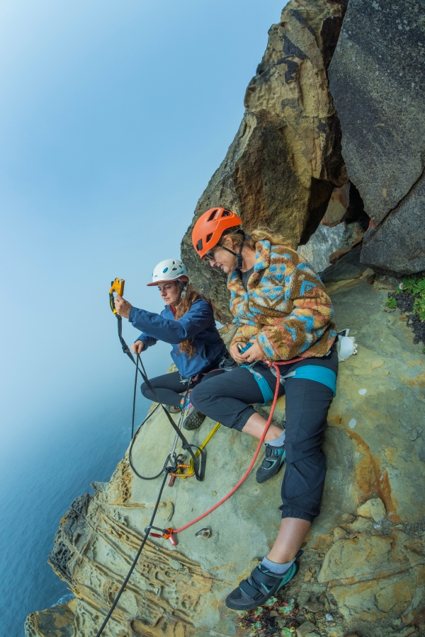 Woman teaching other woman rock climber about using ascenders on sandstone rock cliff overlooking the ocean.