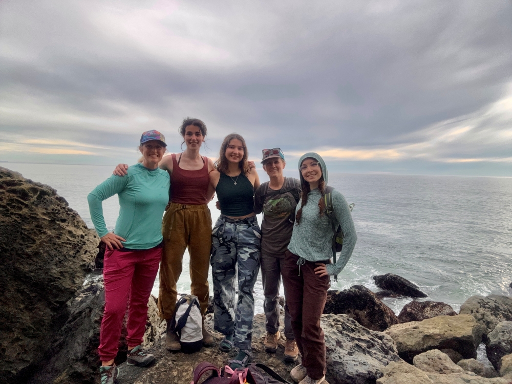 Group portrait of 5 women rock climbers posed in front of the ocean.