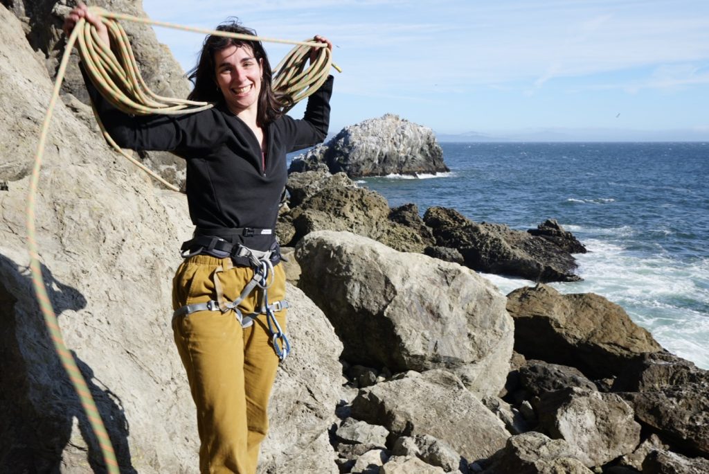 Woman rock climber coiling a climbing rope on rocks near the ocean.